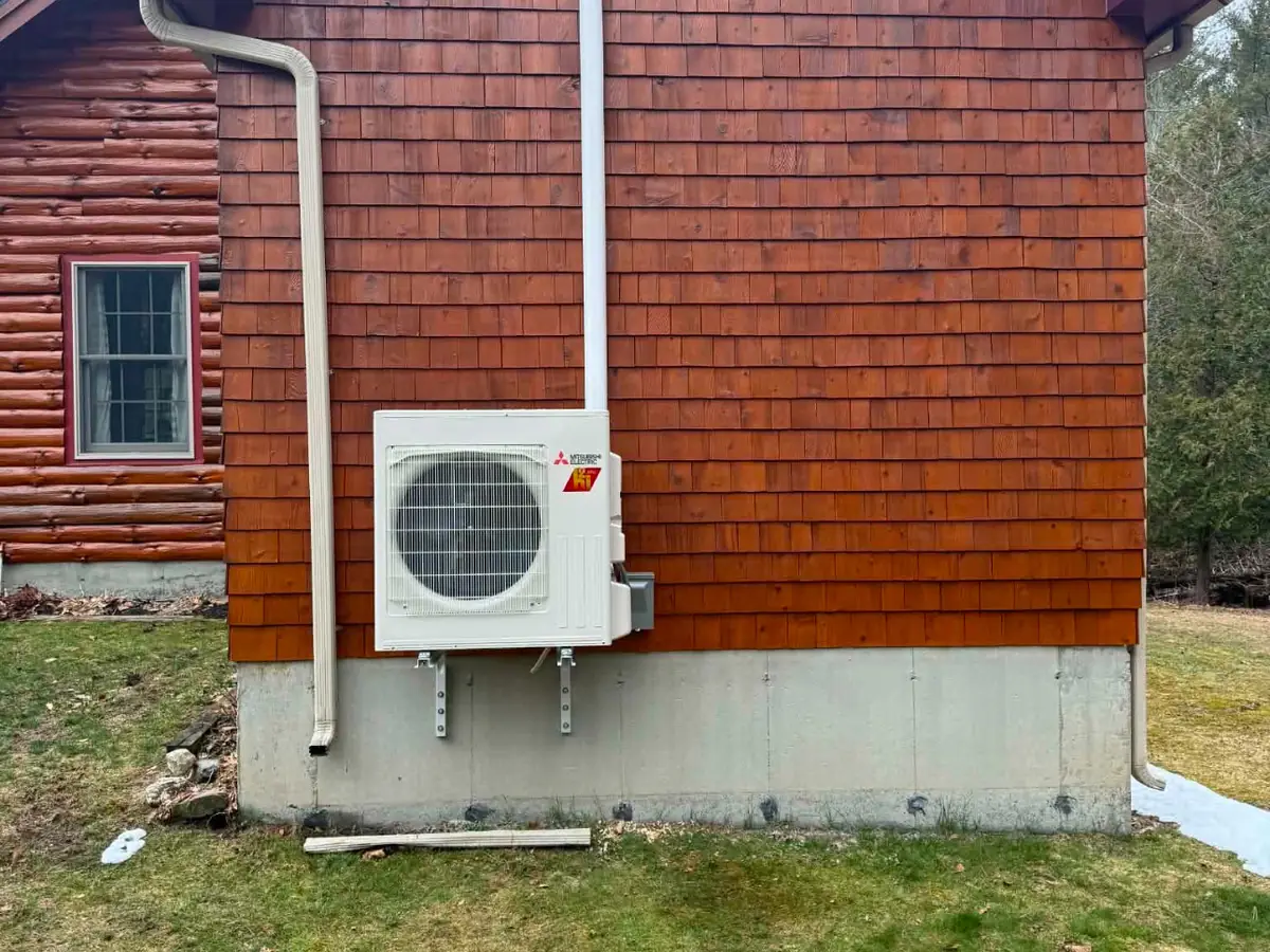 A heat pump outdoor unit mounted on the side of a red shingled house.